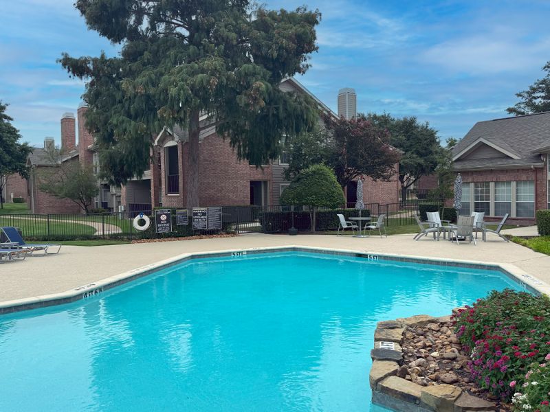 Outdoor pool surrounded by lounge chairs and greenery, with brick apartments in the background under a cloudy sky.