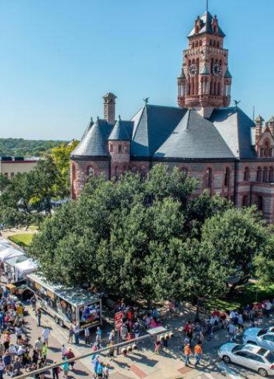 Aerial view of a busy outdoor festival near a historical building with a clock tower and many tents and people.