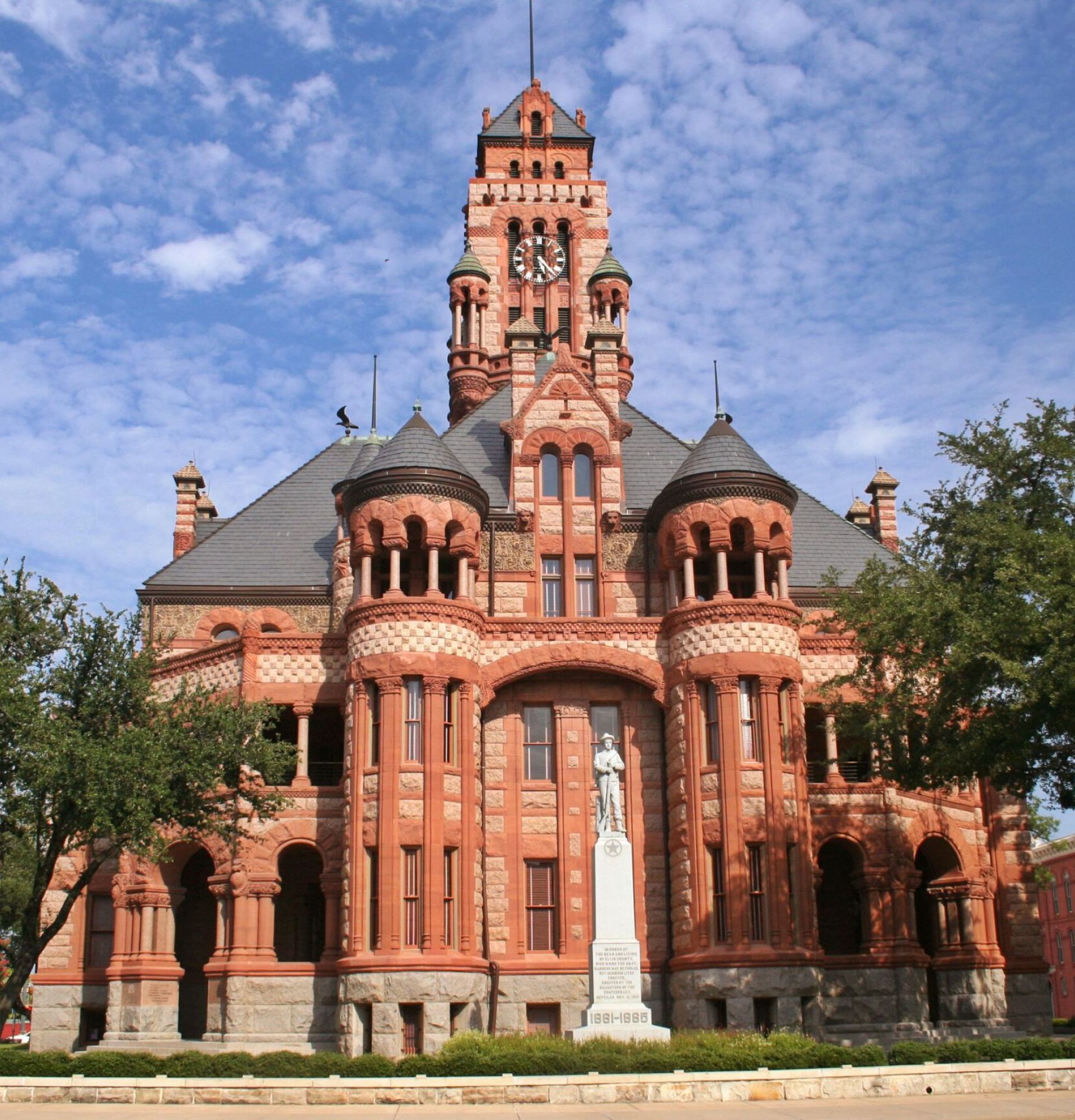 Historic red brick courthouse with a clock tower and statue, surrounded by trees under a partly cloudy sky.