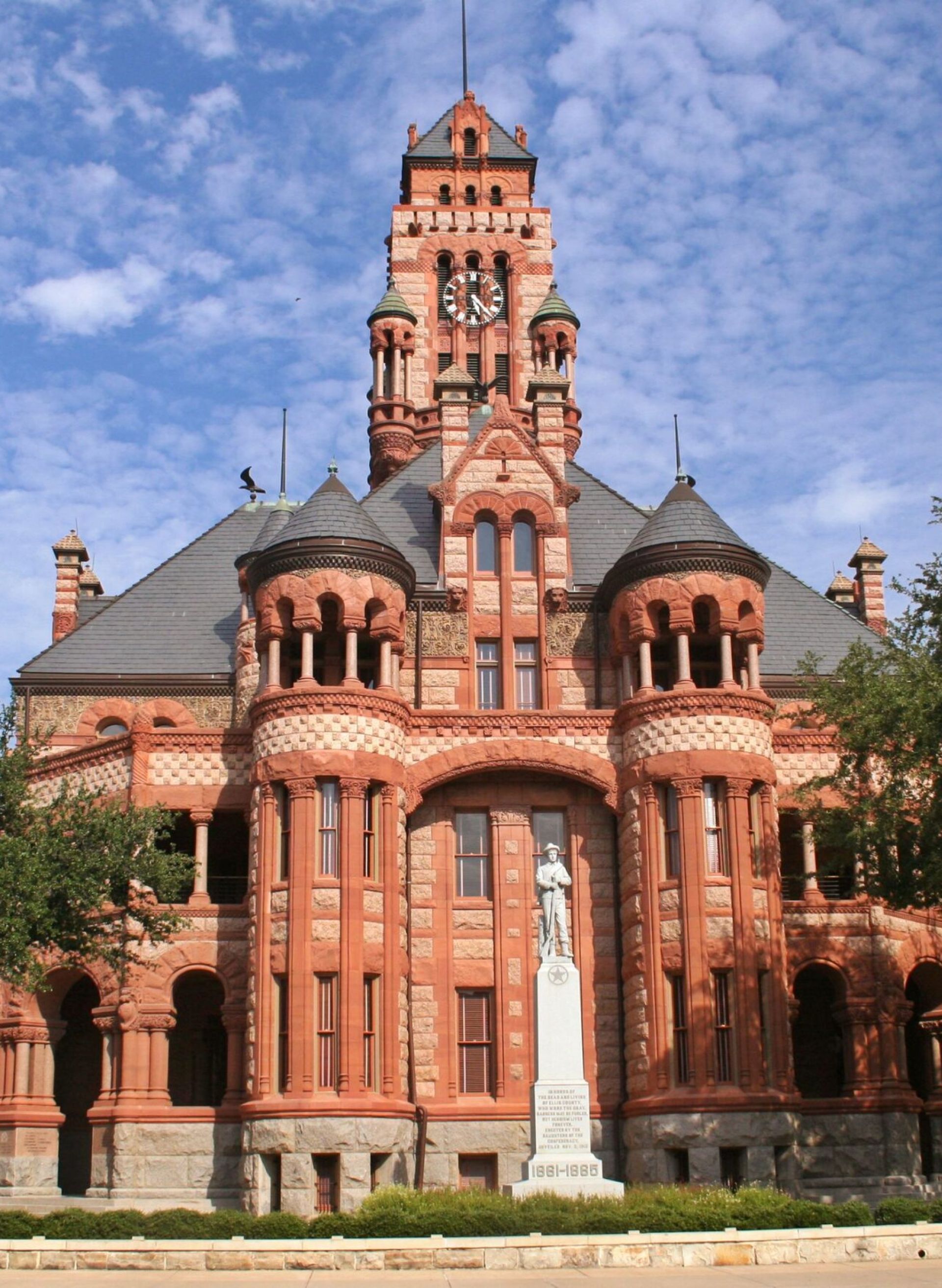 Historic red brick courthouse with a clock tower and statue, surrounded by trees under a partly cloudy sky.