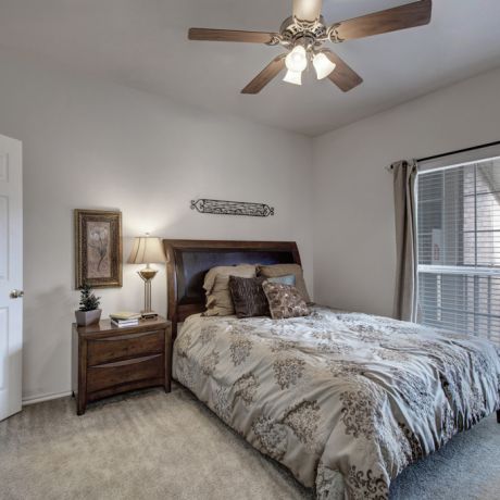 Cozy bedroom with a bed, nightstand, lamp, and ceiling fan, featuring large windows with curtains.