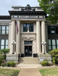 Historic library building with Greek columns, central entrance, and a U.S. flag. Lush greenery in the foreground.