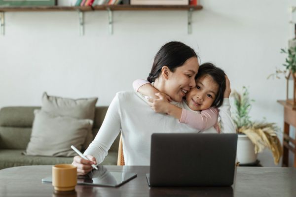 A mother works on a tablet at a table, smiling while her child hugs her from behind, with a laptop in front.