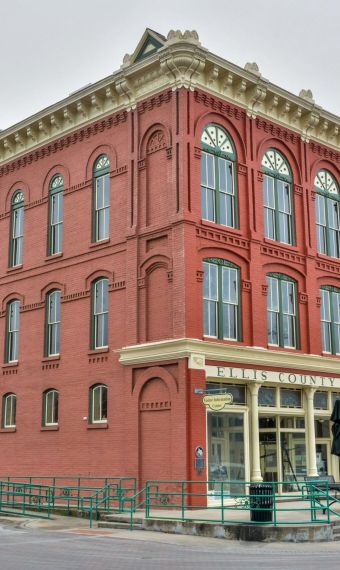 Historic red brick building housing Ellis County Museum, with arched windows and parked cars in front.