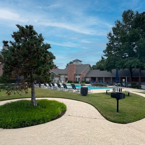 Community pool surrounded by lounge chairs, greenery, and trees, with residential buildings in the background.